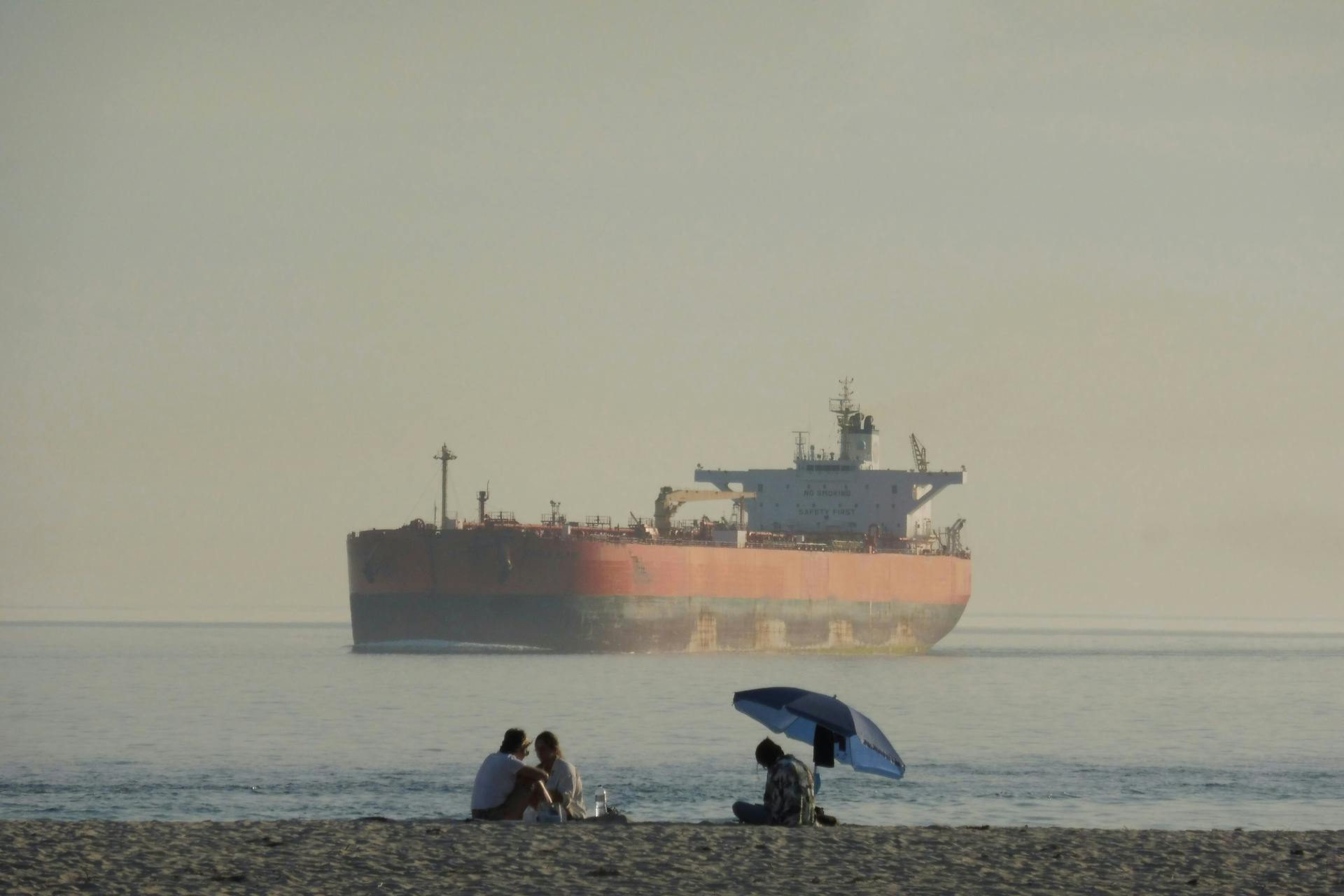 Oil tanker anchored near a beach where people relax under an umbrella.
