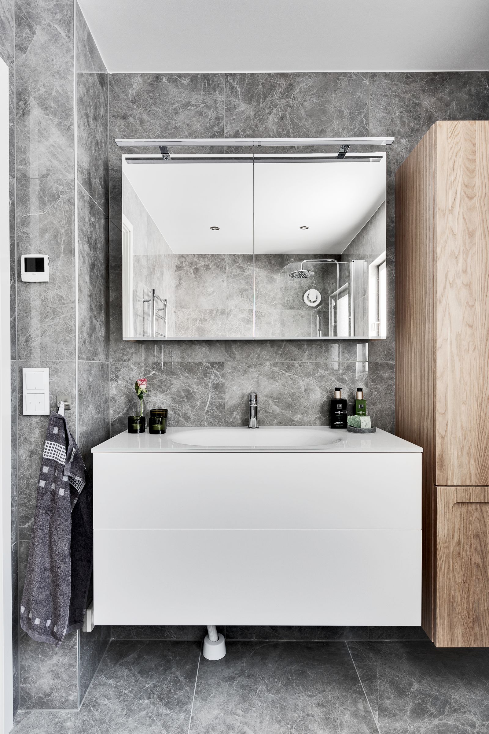 Modern bathroom vanity with a white floating cabinet, integrated basin, large mirrored cabinet, grey marble-look wall and floor tiles, and wood-toned storage.