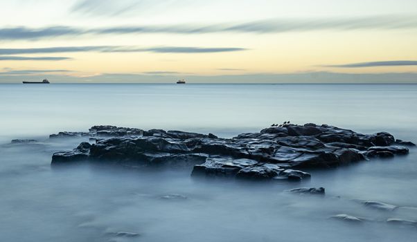 A tranquil seascape with a rocky shoreline and distant ships at twilight, capturing the calmness of the ocean.