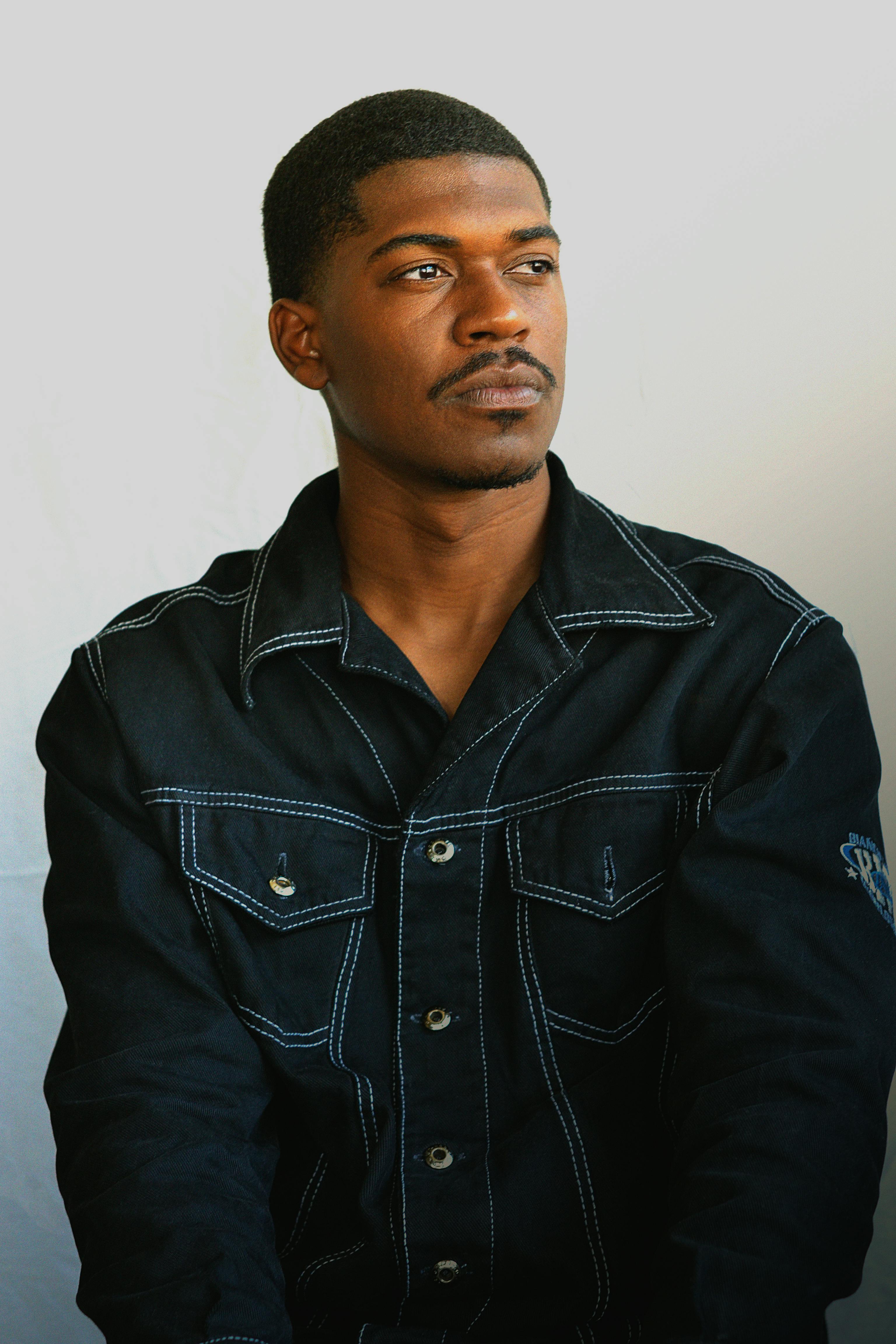 A thoughtful young man in a denim jacket looks to the side in a studio setting.
