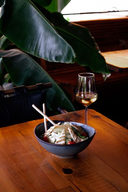 A bowl of noodles with chopsticks and wine on a wooden table, indoor setting.