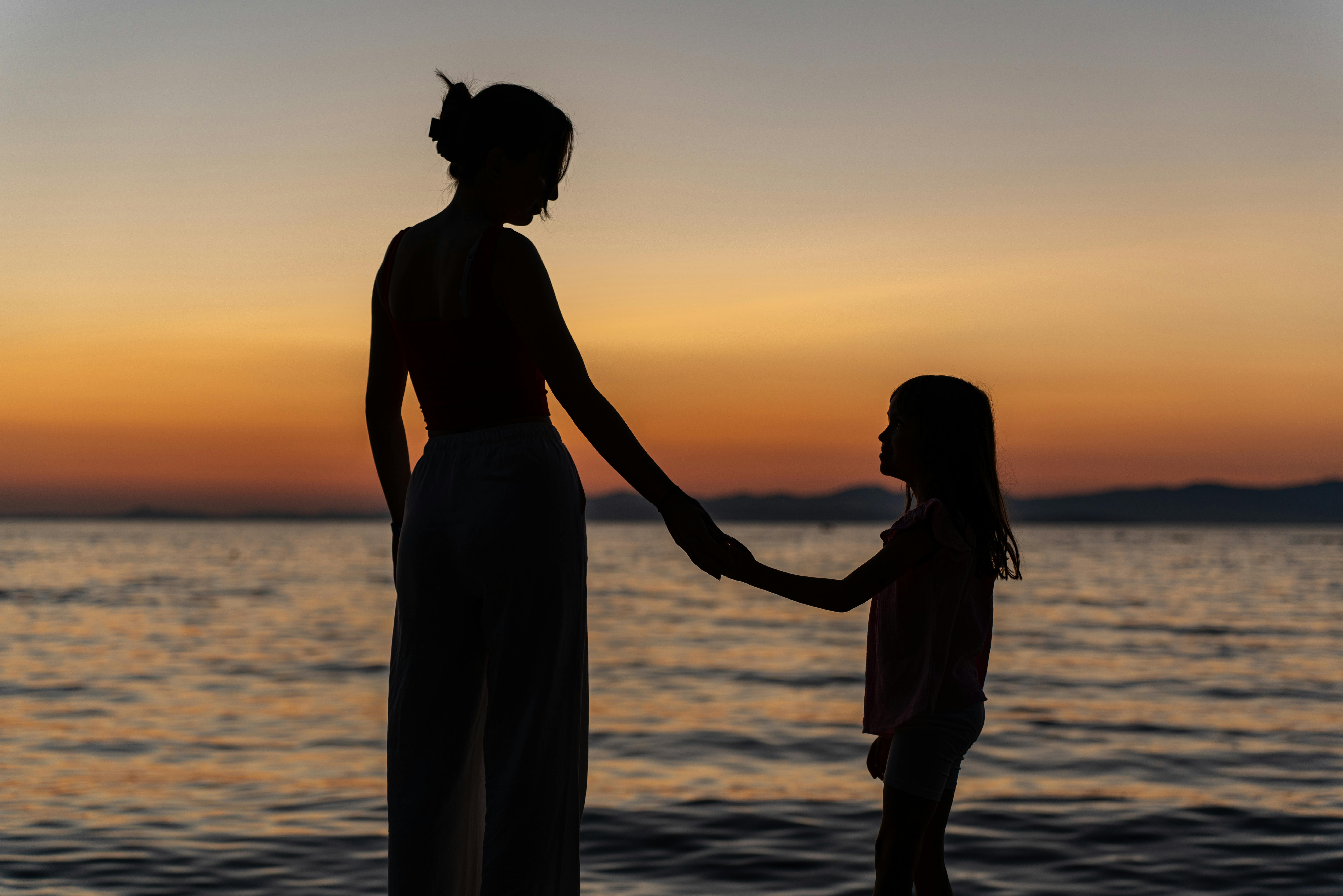 A serene silhouette of a mother and daughter holding hands by the ocean during sunset, symbolizing love and connection.