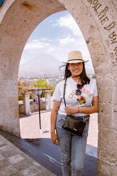 A woman wearing casual attire smiles under a historic stone archway with a scenic mountain view in Arequipa, Peru.