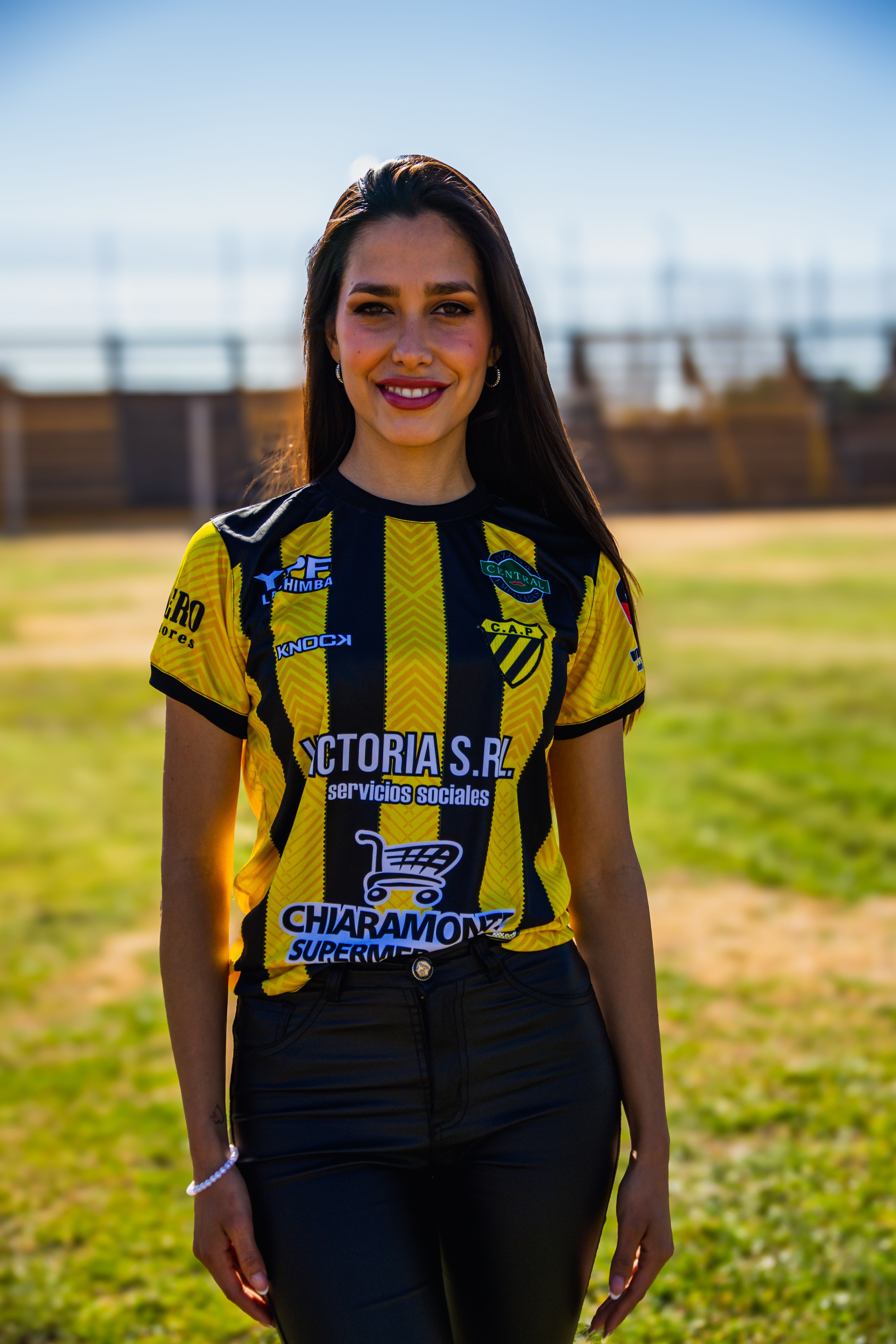 Smiling woman in a black and yellow sports jersey standing on a sunny field.