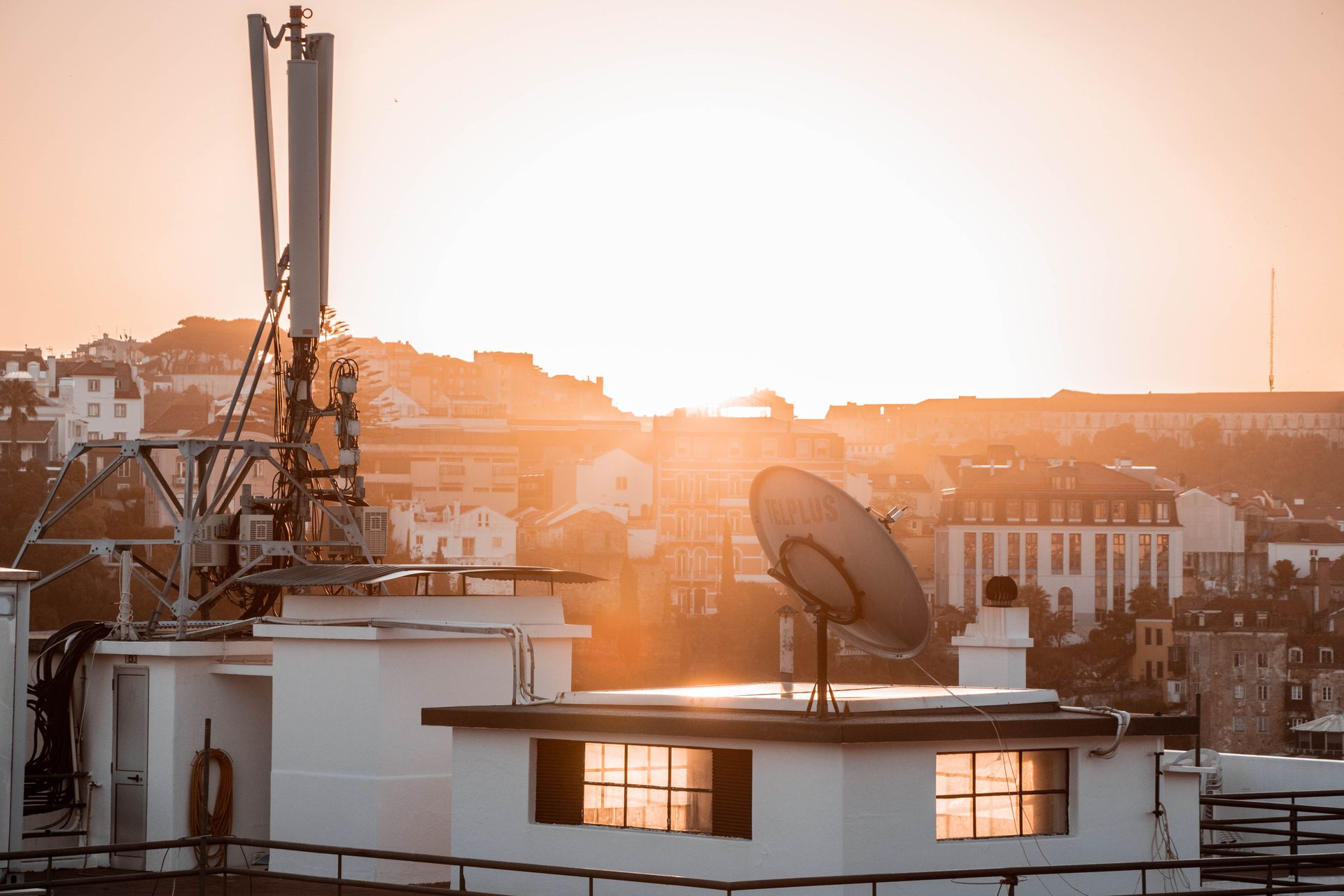 A cityscape view of rooftops and satellite dish illuminated by the rising sun.
