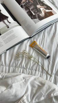 A stylish open magazine with a skincare tube and delicate flower on a white bedspread.