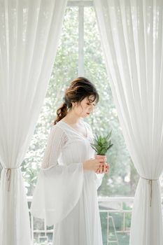 A serene portrait of a woman in a white dress holding a potted plant, framed by sheer curtains.