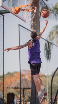 A young man jumps to dunk a basketball on an outdoor court during daytime play.