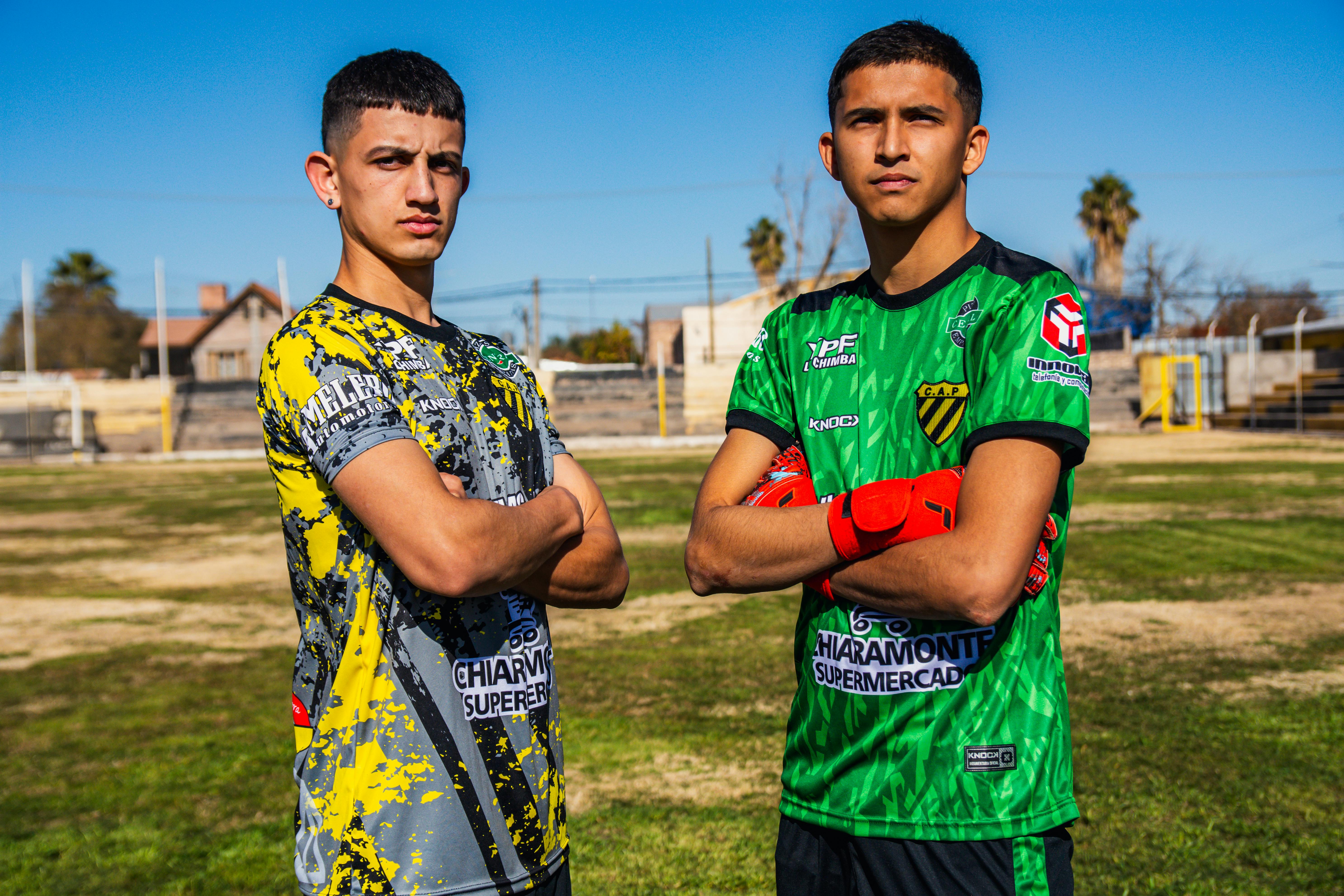 Two young men proudly posing on a soccer field, wearing team jerseys.