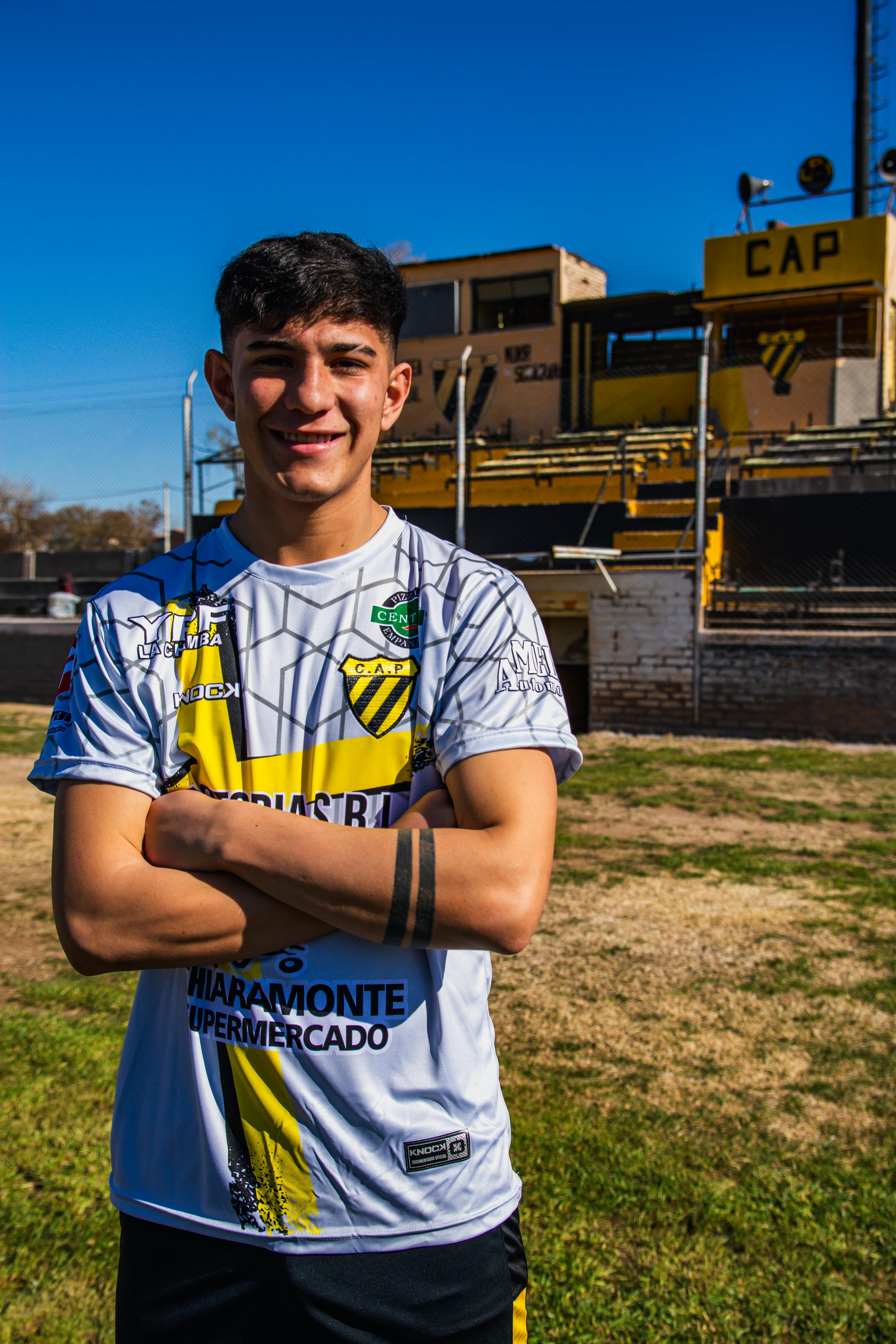 Smiling young man in a sports jersey stands confidently on a football field under a clear blue sky.