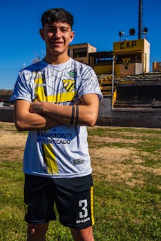 Confident young male soccer player posing outdoors on a sunny day with stadium background.