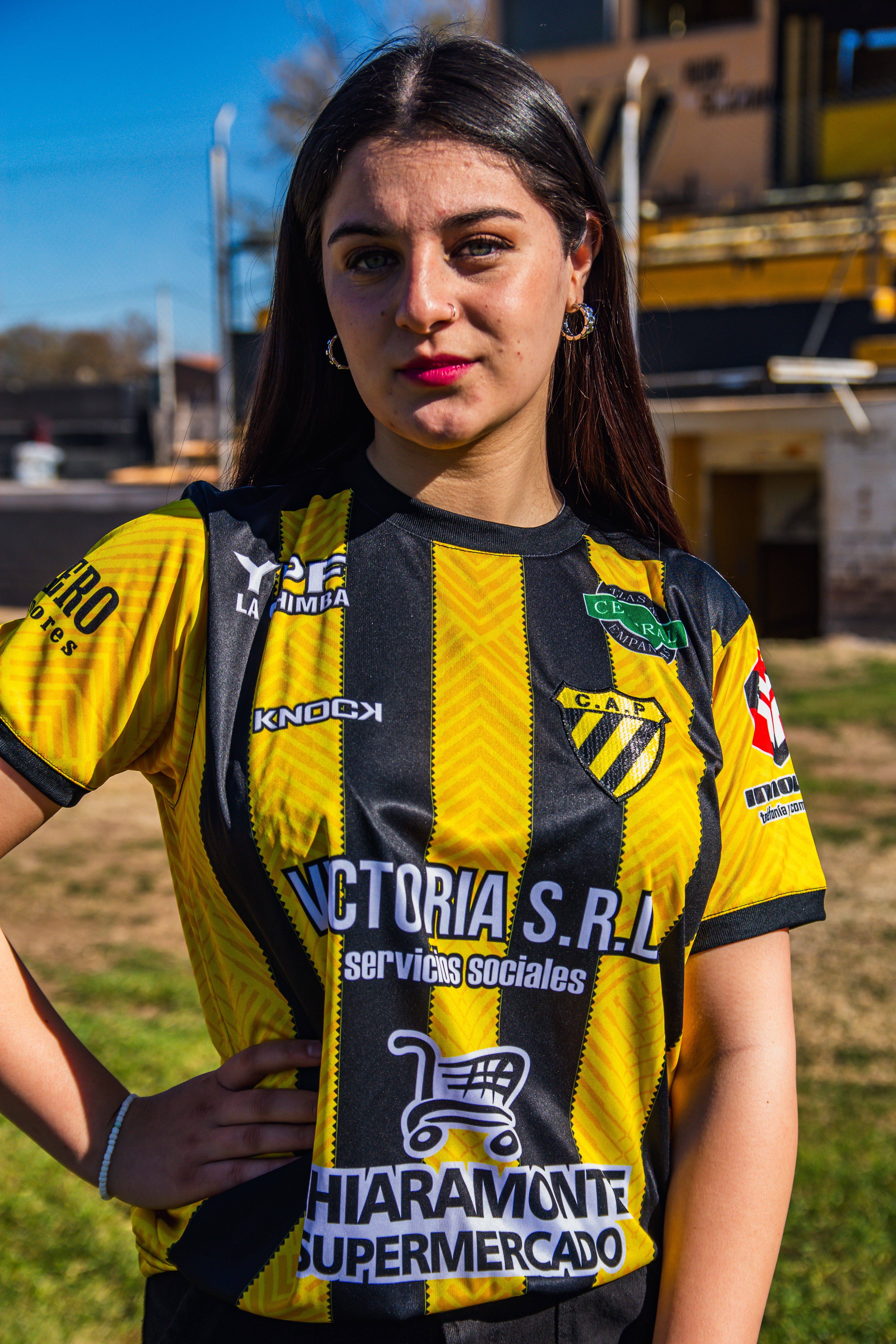 Young woman proudly wearing a black and yellow soccer jersey, standing outdoors on a sunny day.
