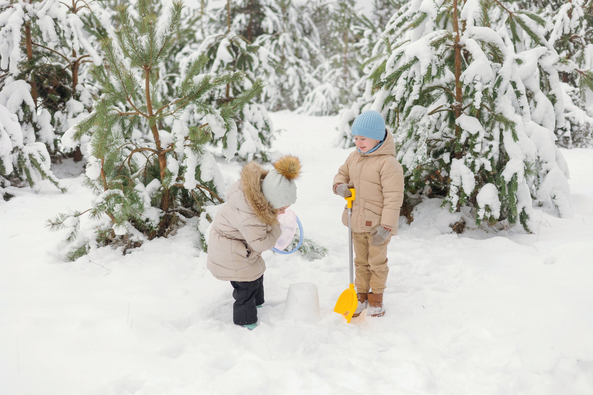 Two children enjoy a snowy day, playing with a shovel in a winter forest.