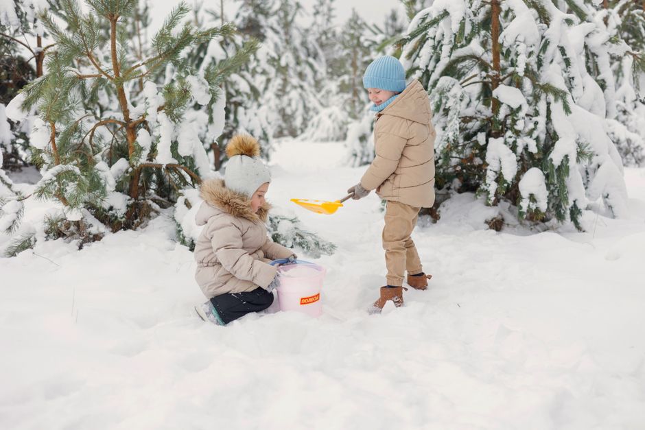 Two kids playing with buckets and shovels in a snowy forest setting, enjoying winter fun.