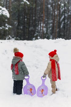 Two children in winter clothing ready to sled on a snow-covered hill.