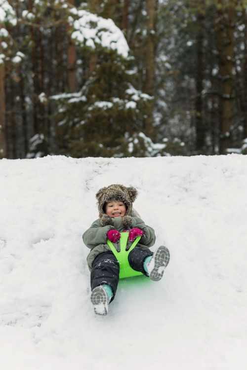 Free A cheerful child sleds down a snowy hill in a winter forest, enjoying the season. Stock Photo