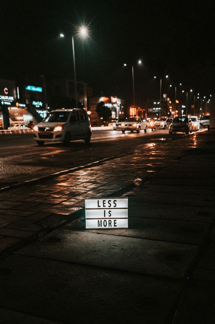 A nighttime street view featuring cars and an illuminated sign on the sidewalk displaying 'Less is More'.