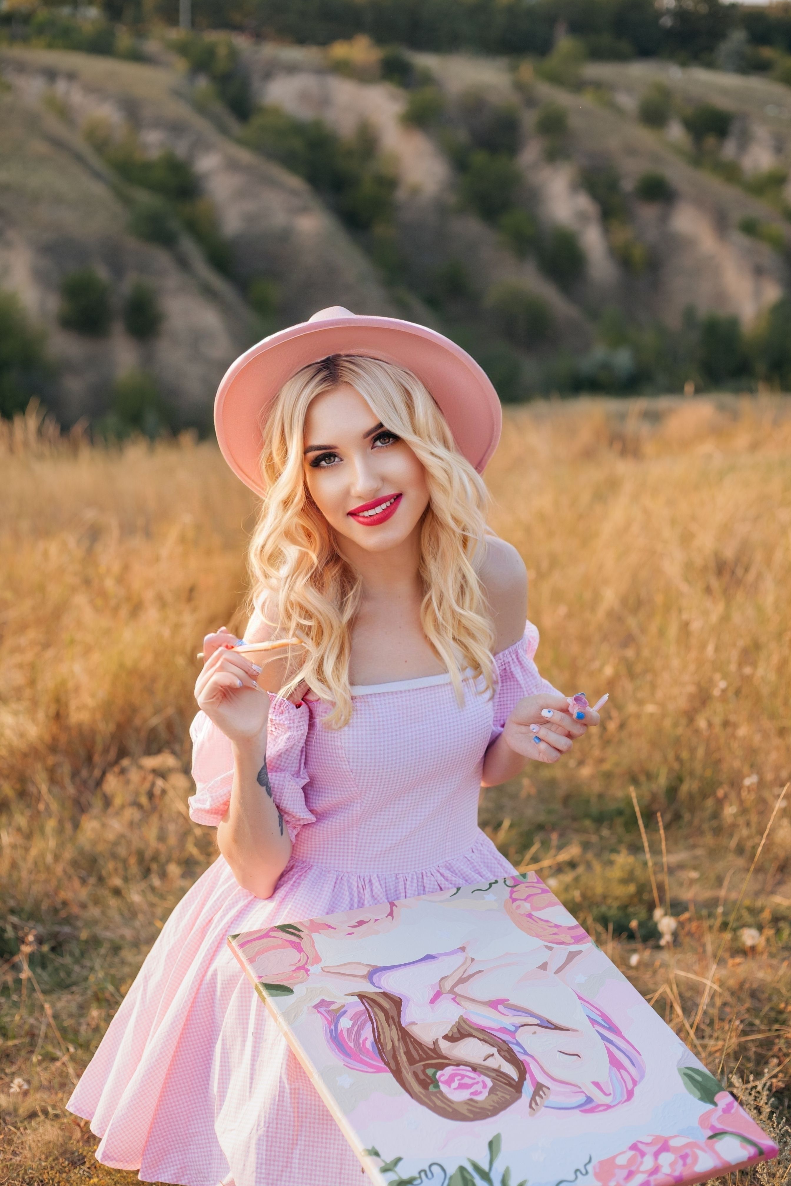 A smiling young woman in a pink dress paints a portrait in a sunny landscape, enjoying a day in nature.
