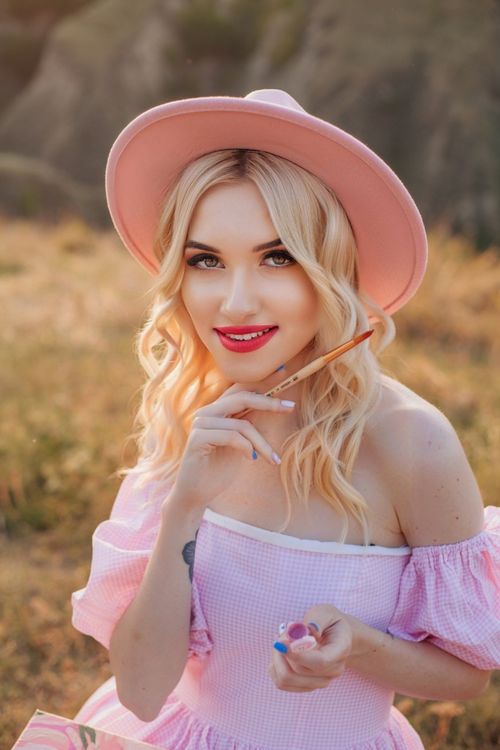 Free Blonde woman in pink dress and hat enjoying a sunny day outdoors with a smile. Stock Photo