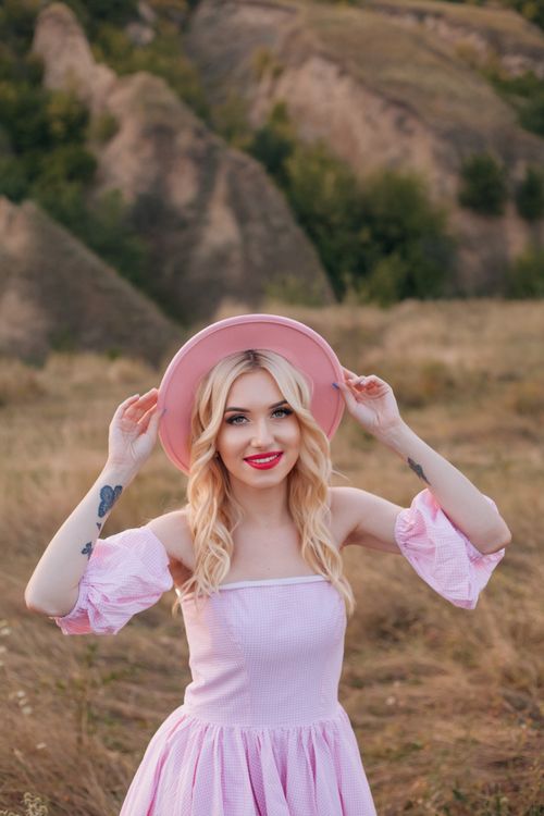 Free Blonde woman in a pink dress and hat enjoying a scenic outdoor setting during sunset. Stock Photo