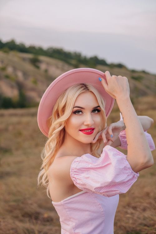 Free Blonde woman with pink hat smiles in a scenic outdoor summer setting. Stock Photo