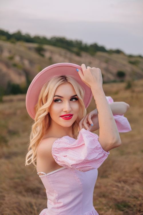 Free Blonde woman in a pink dress with a hat, smiling in a picturesque outdoor setting during sunset. Stock Photo