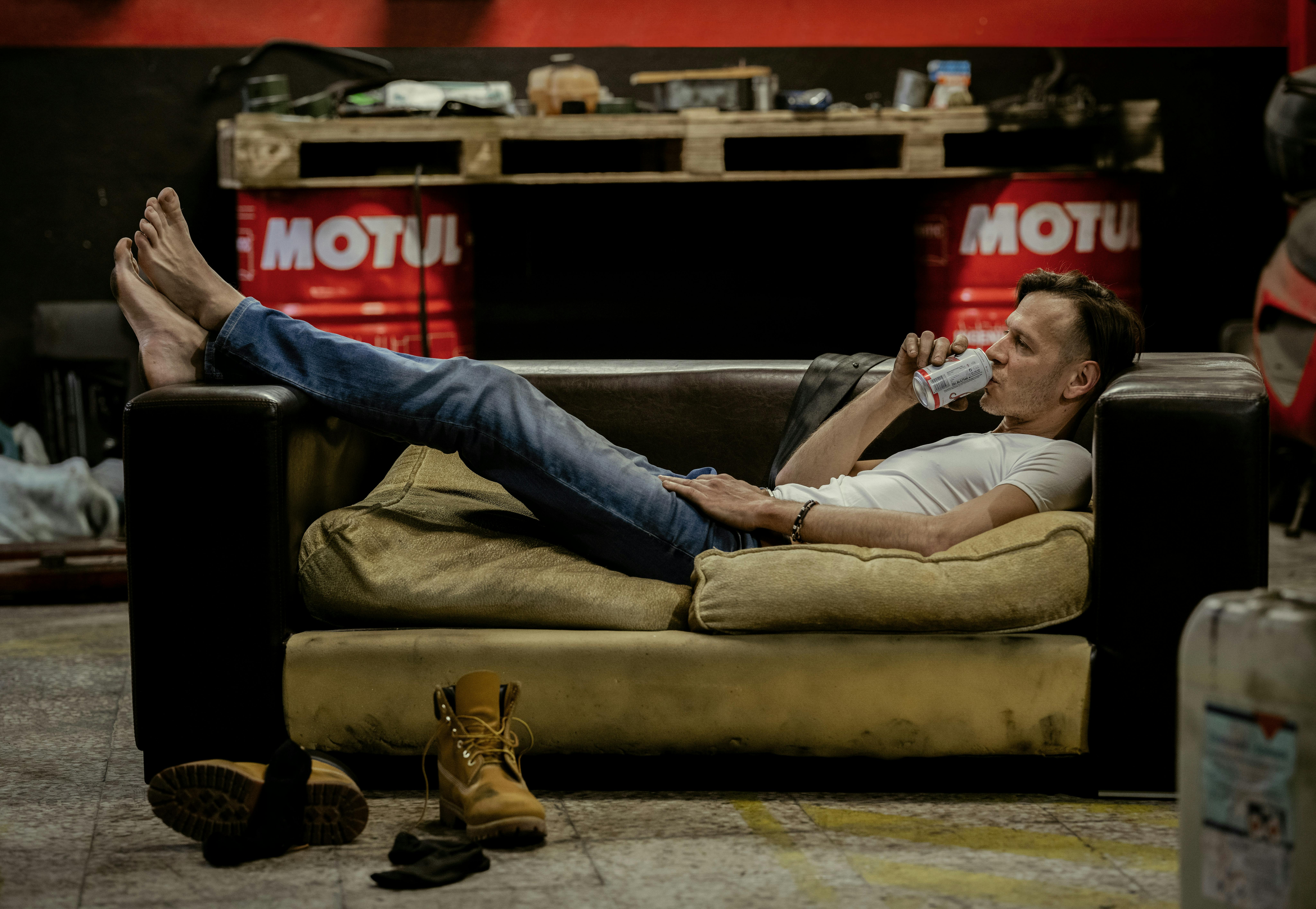 Man lounging on a sofa in a garage, drinking a beverage, surrounded by tools and equipment.