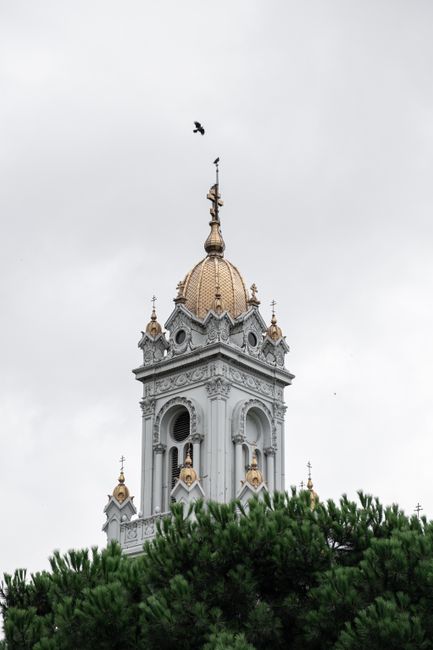 A church tower with a golden dome set against a cloudy sky, featuring intricate architecture above green trees.