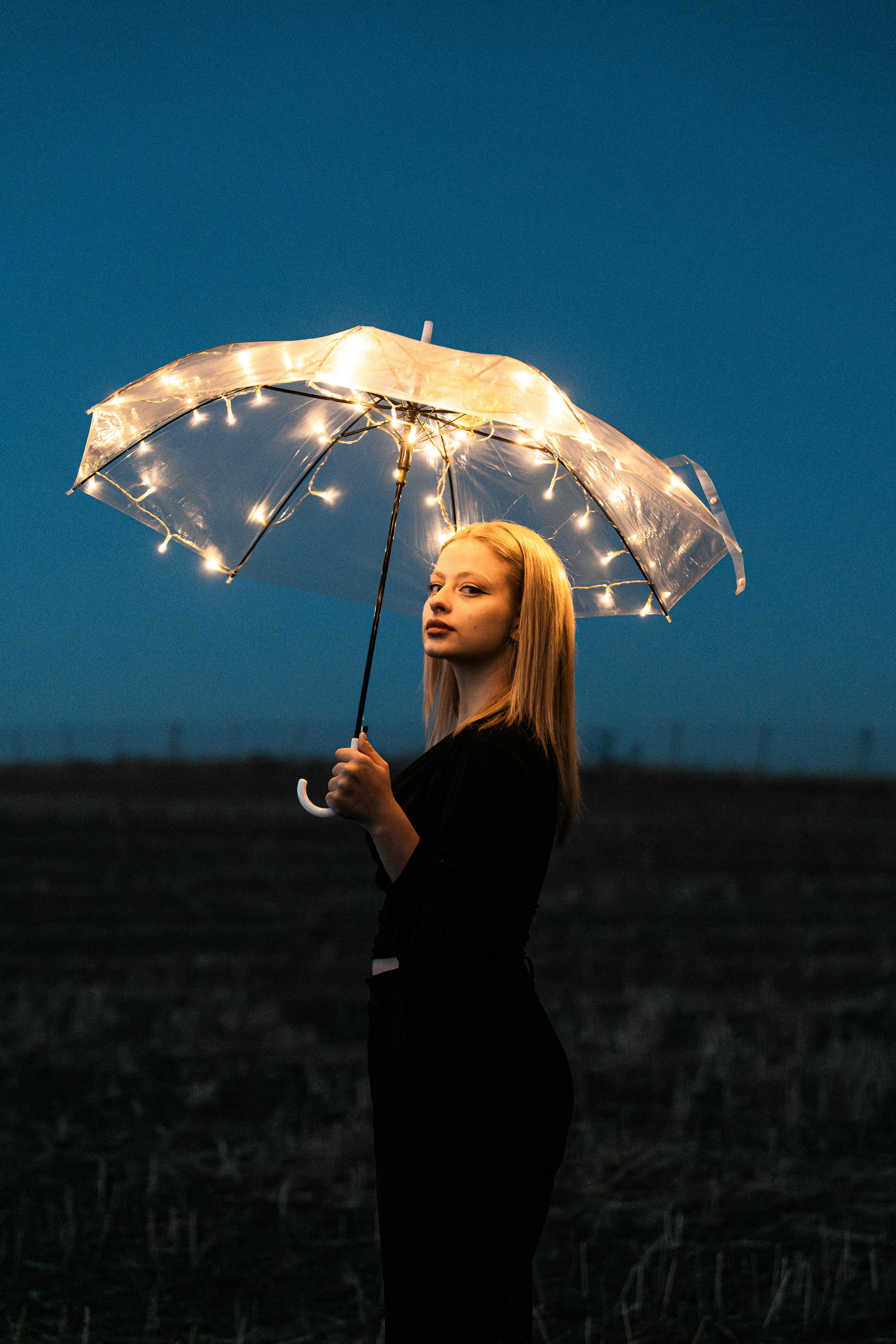 A woman with a glowing umbrella stands in a rural field, creating an ethereal nighttime scene.