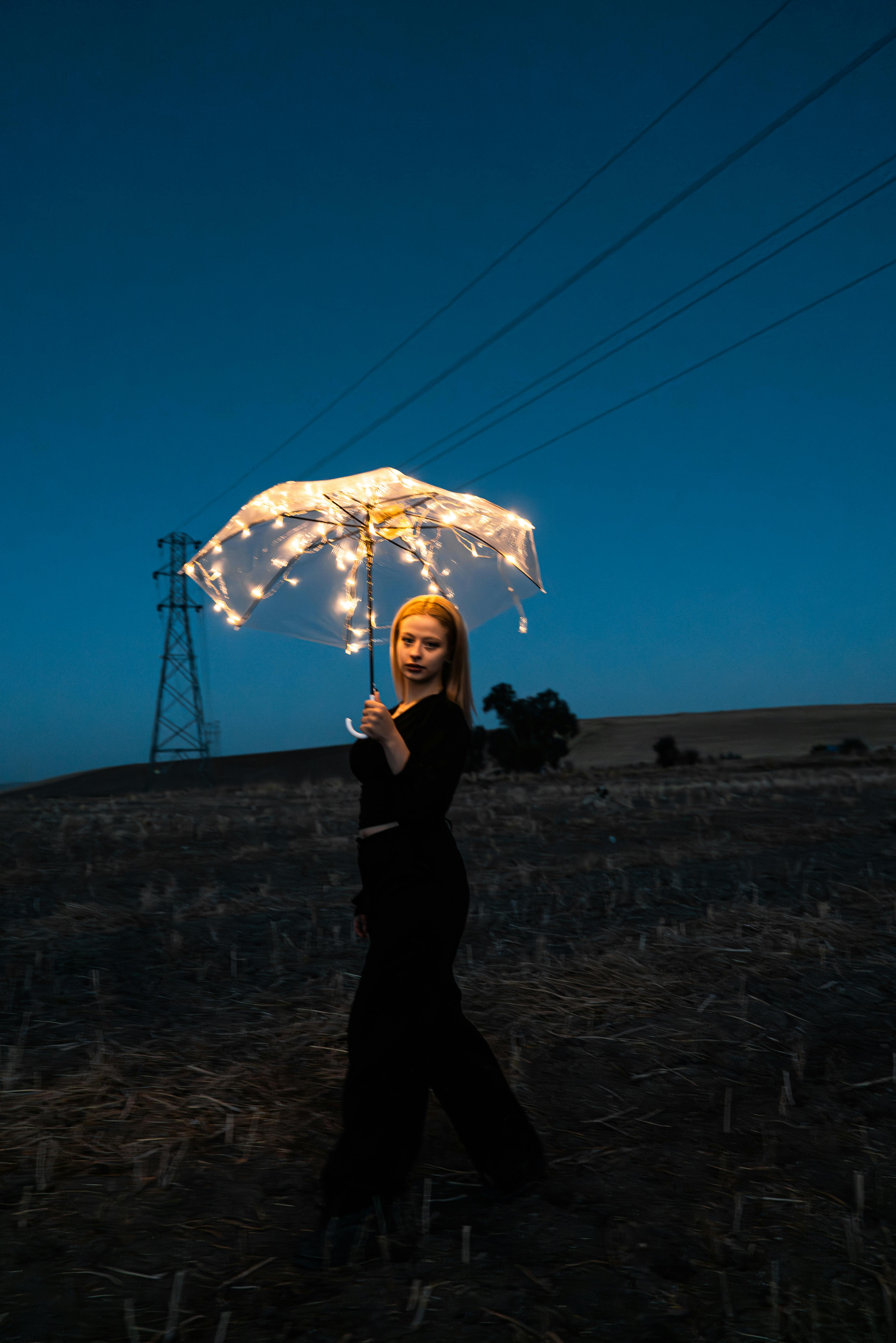 A woman stands in a field holding a fairy light-adorned umbrella at dusk, creating a magical ambiance in Ankara, Türkiye.