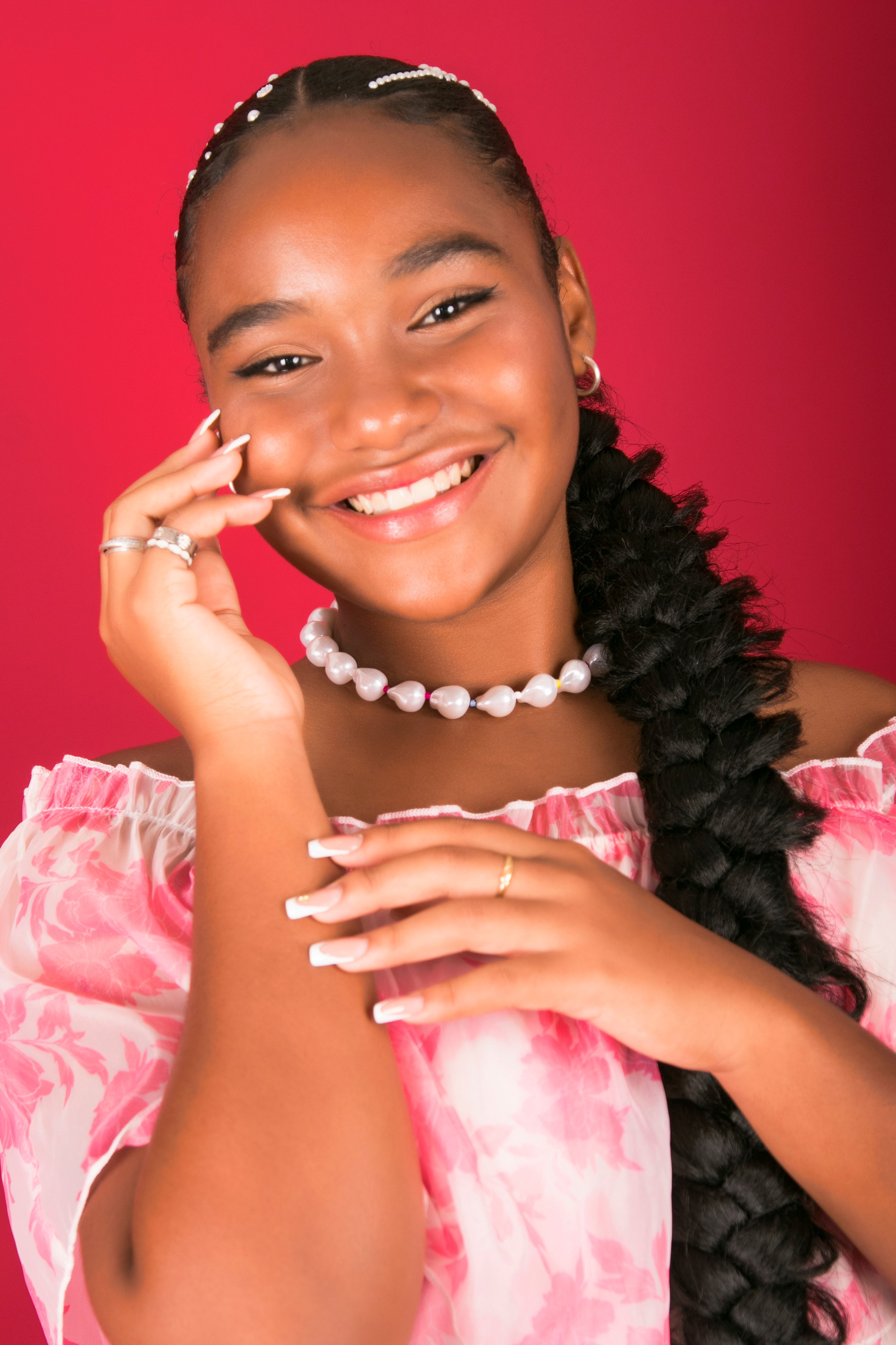 Portrait of a smiling young woman with braids and pearls, against a vibrant red background.