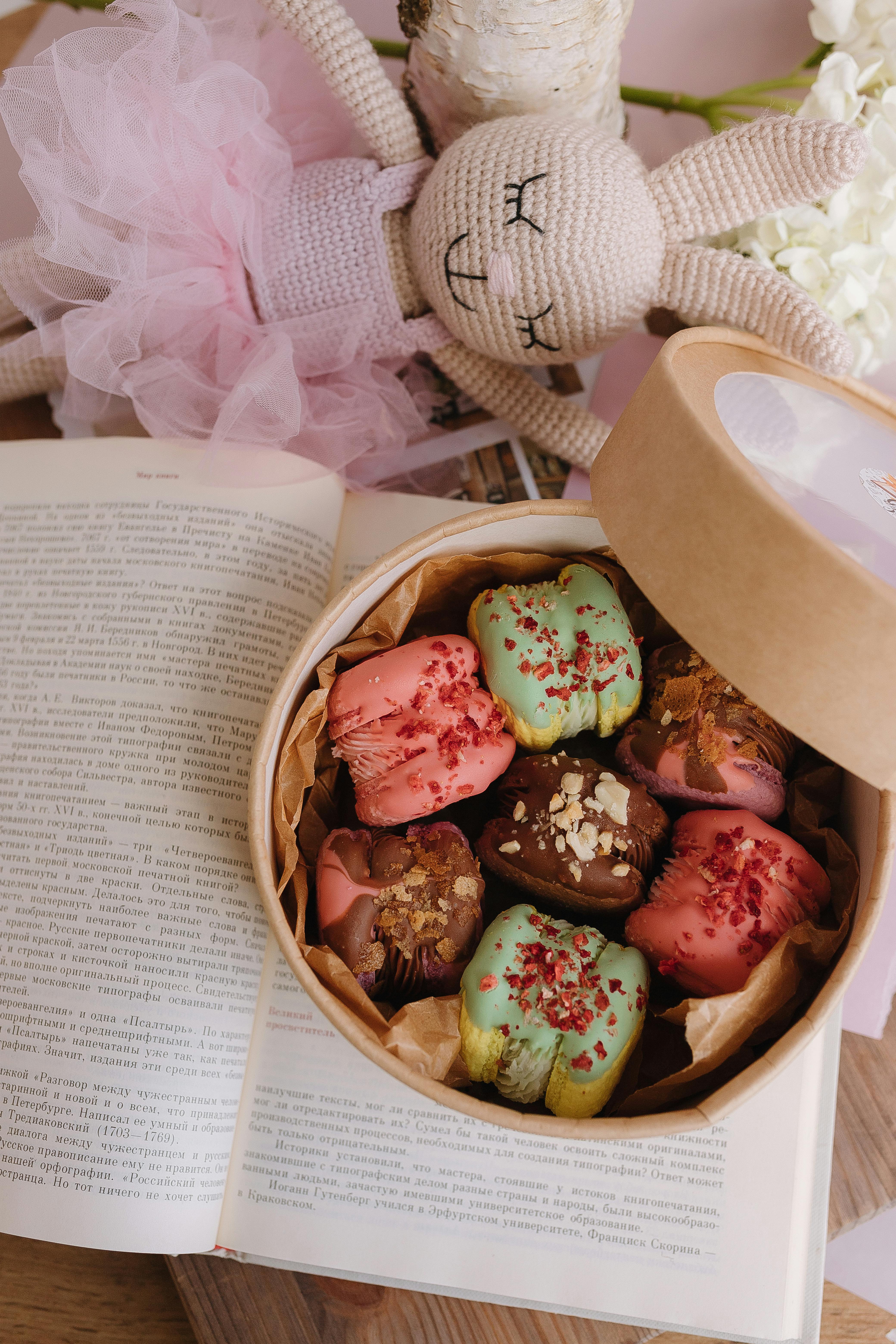 Elegant box of decorated cupcakes beside a handmade crochet doll in a cozy indoor setting.
