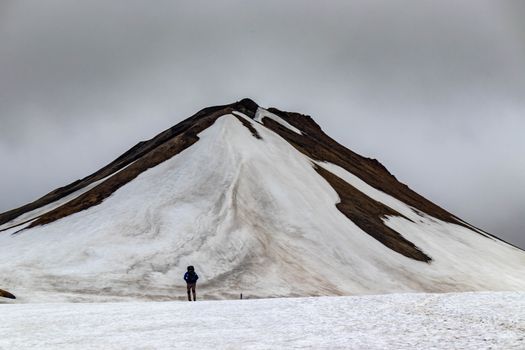 A solitary hiker stands before a striking, snow-covered mountain in Iceland, showcasing the rugged wilderness.