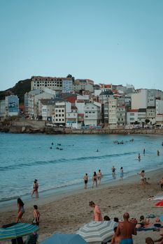 People enjoying a sunny day at the beach in Malpica, Spain, with a scenic coastal town backdrop.