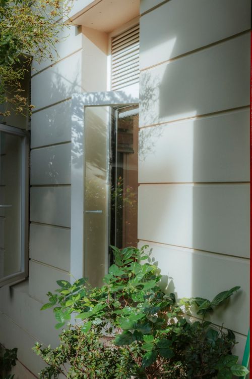 Free Sunlit modern home exterior with open window and vibrant greenery, creating a serene summer ambience. Stock Photo