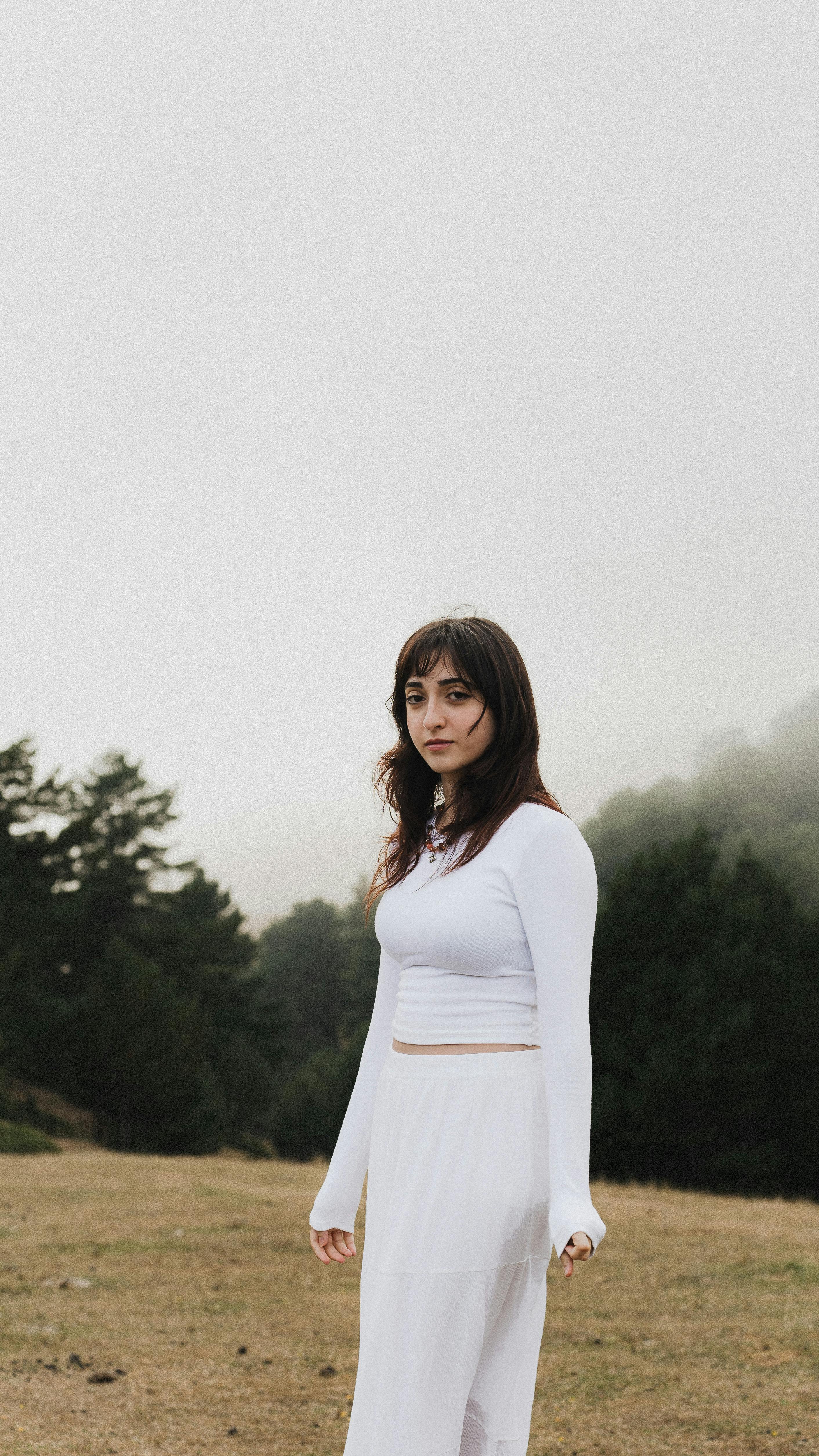 A woman in a white dress standing in a misty, serene outdoor setting with trees.