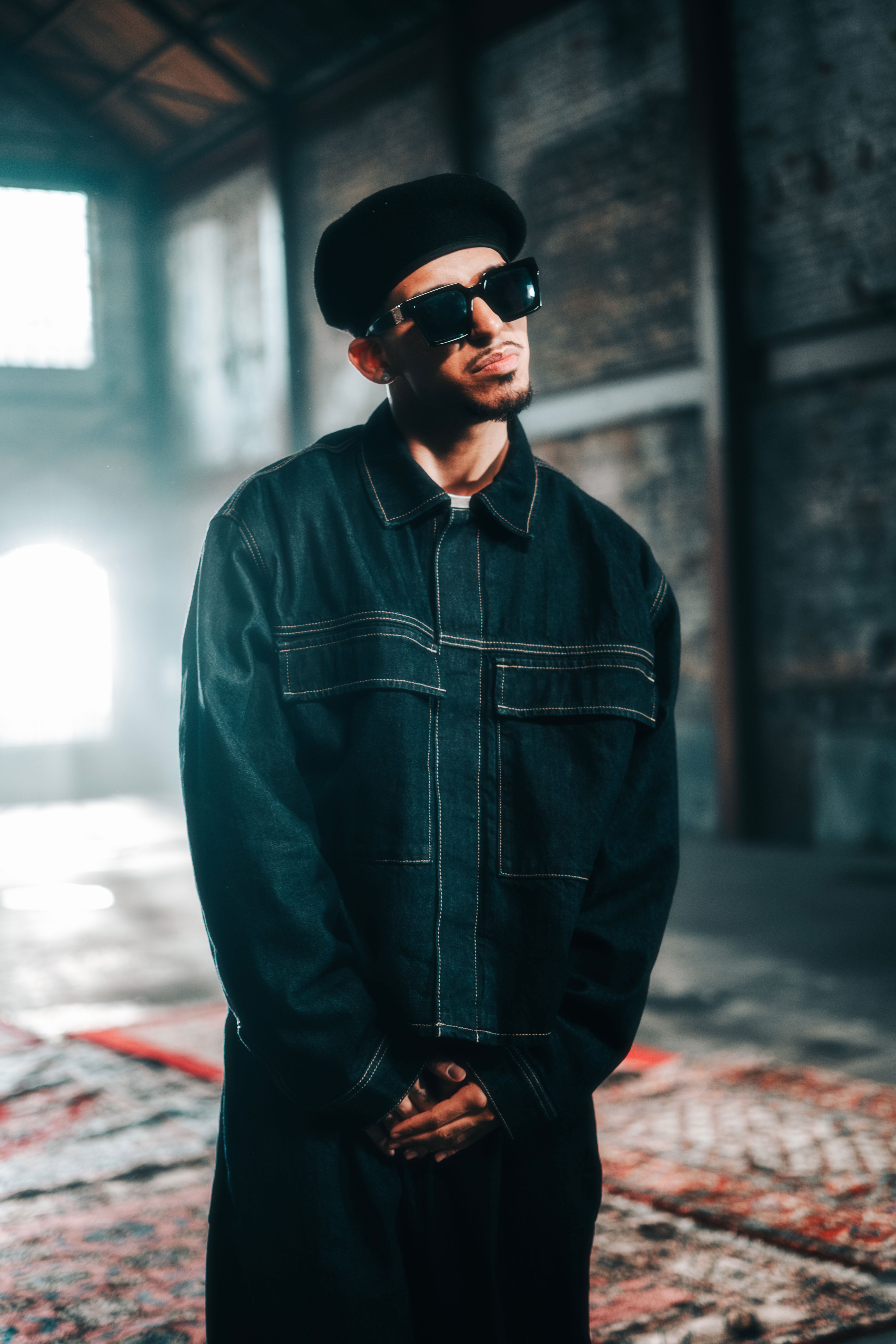 Fashionable man with sunglasses in urban industrial setting, wearing trendy outerwear.