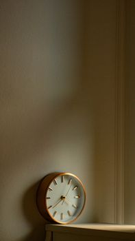 A retro-style analogue clock resting on a wood surface, casting warm light against a white wall.