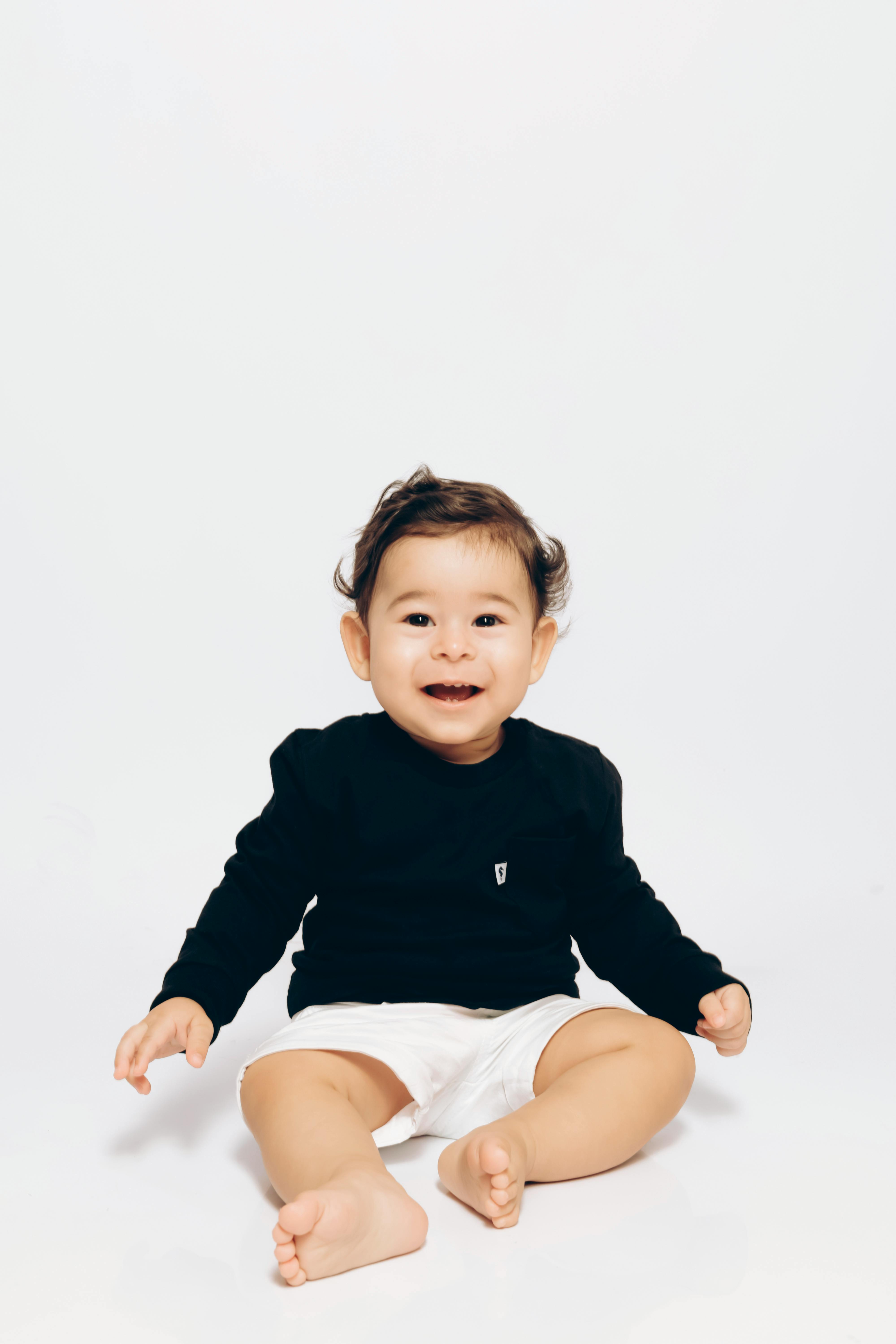 Charming baby in a black shirt and white shorts sitting and smiling in a bright studio setting.