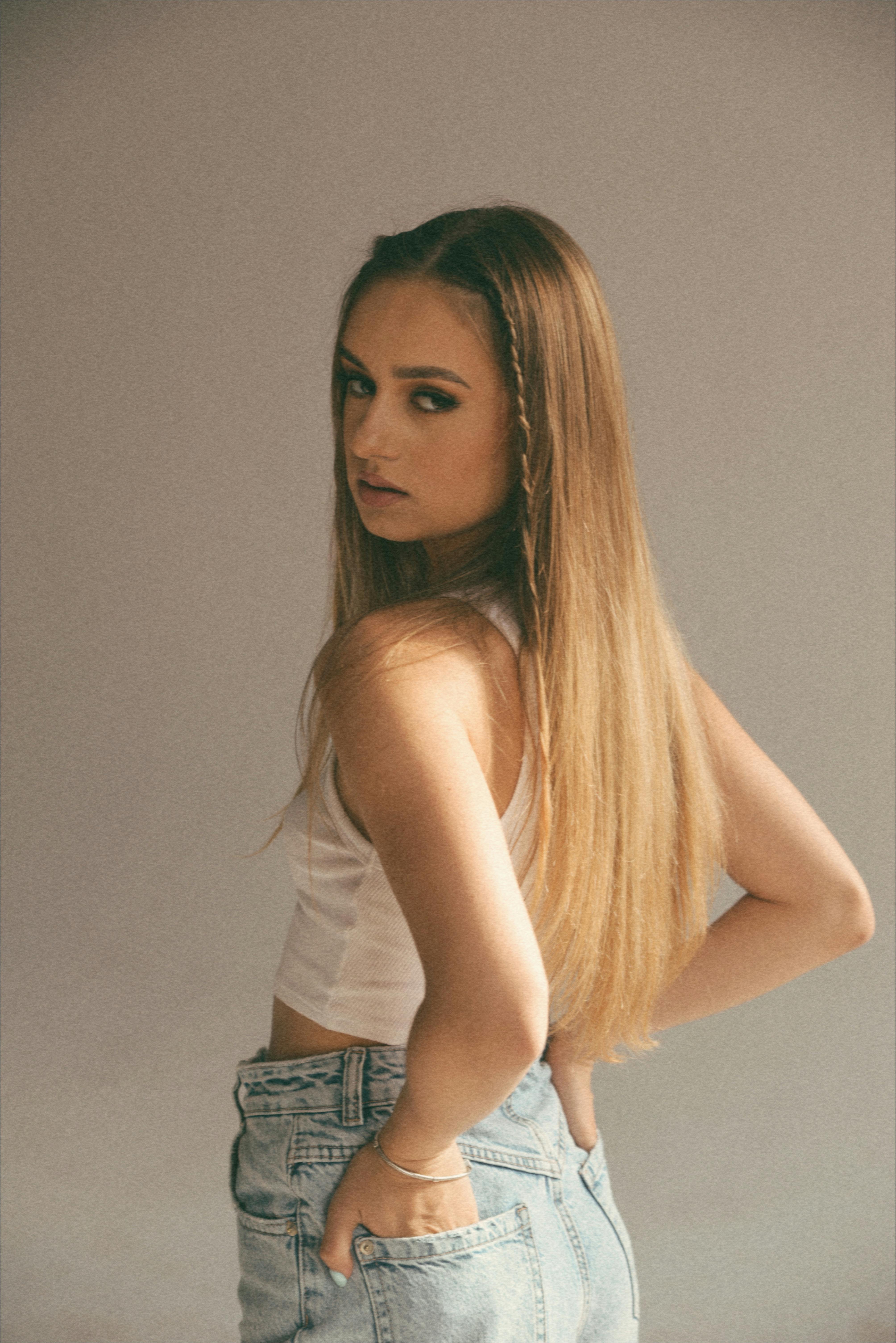 Stylish young adult woman poses indoors wearing denim jeans and a white crop top.