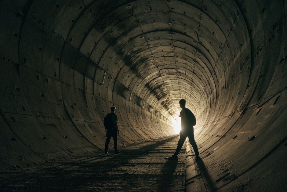 Two people silhouetted in a dramatic, dark industrial tunnel with a light source at the end.