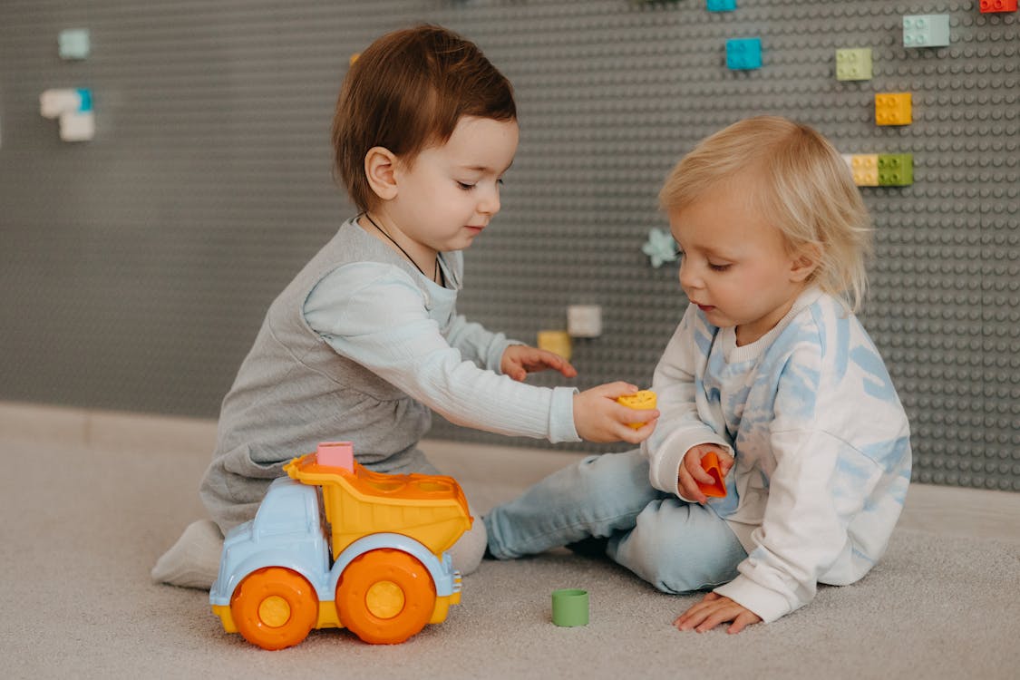 Free Two toddlers happily playing with a toy truck indoors, surrounded by colorful blocks. Stock Photo