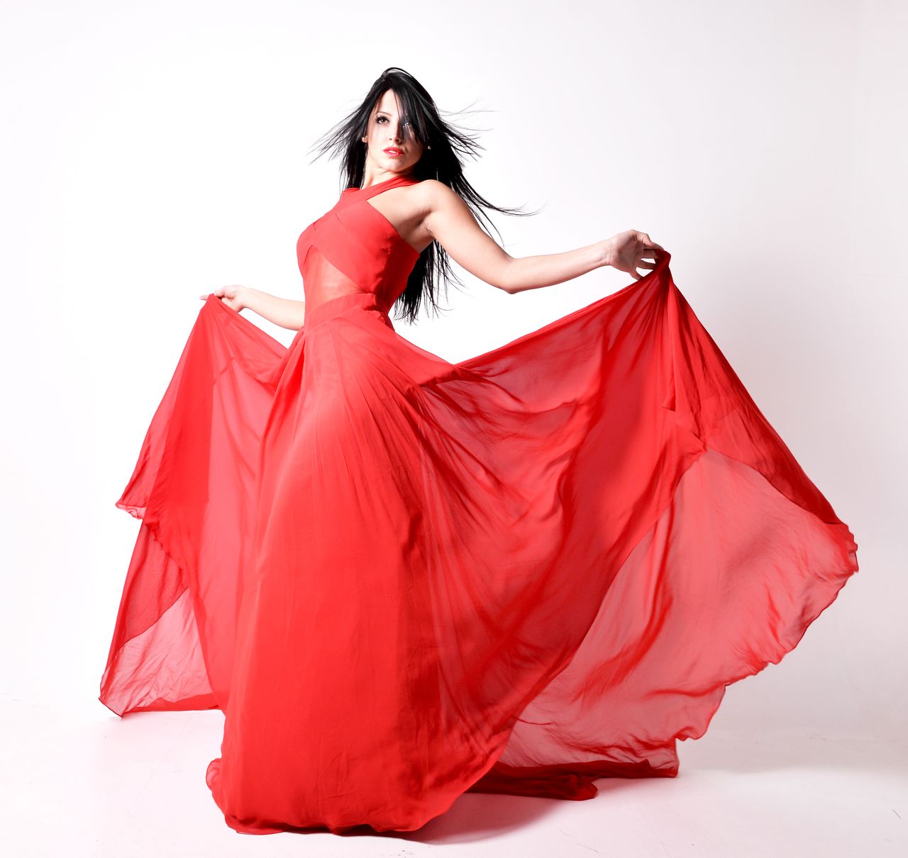 Woman in a flowing red chiffon evening dress holding out the skirt against a white background
