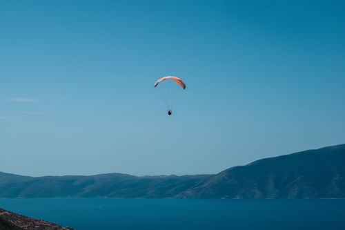 Free Paraglider soaring over tranquil sea with distant mountains under clear blue sky. Stock Photo