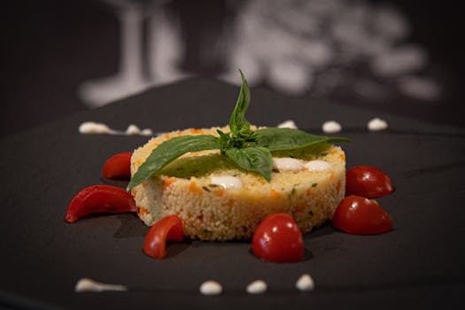 A close-up of a gourmet couscous salad garnished with fresh cherry tomatoes and basil leaves on a dark plate.