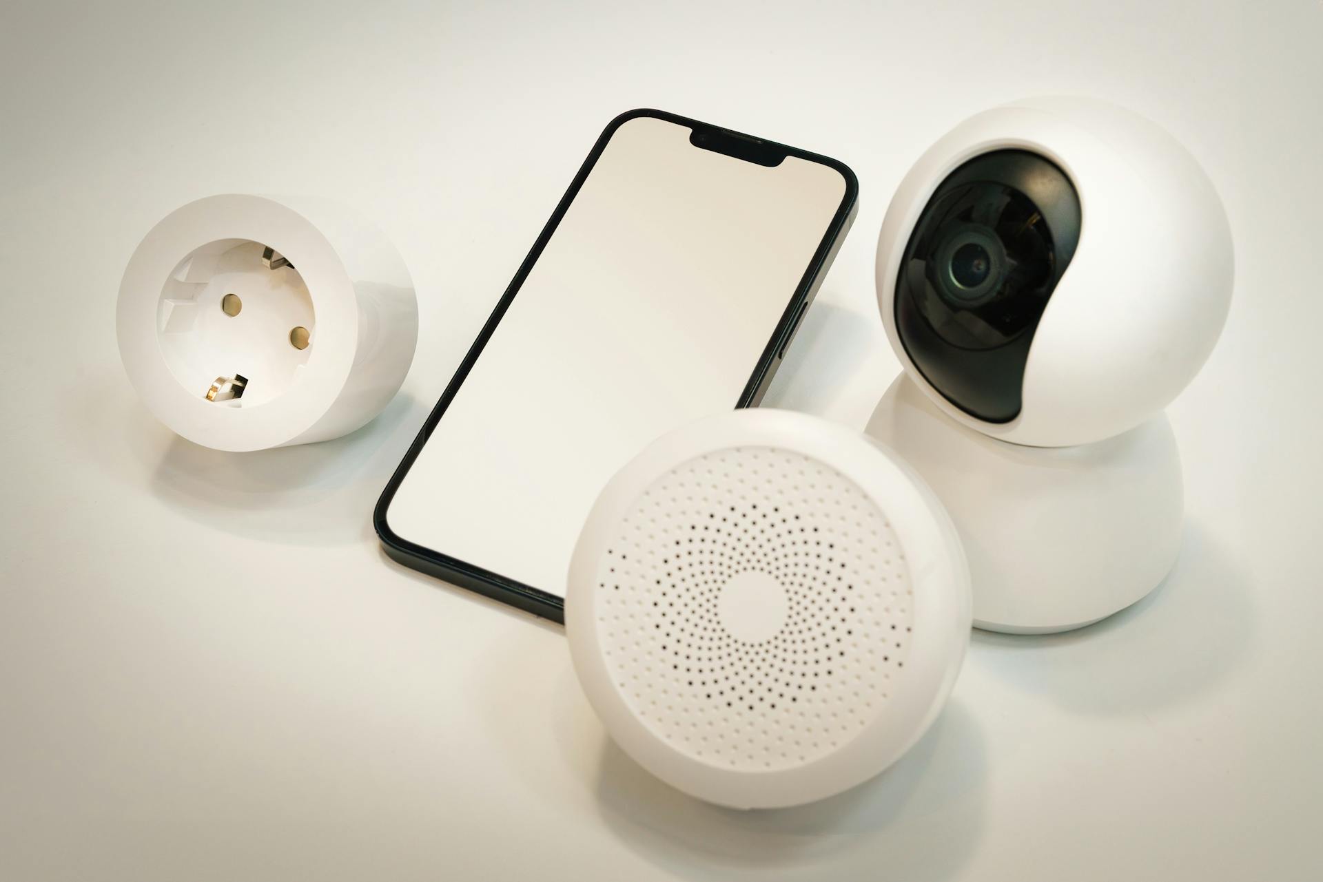 Smart home gadgets including a security camera, speaker, and smartphone displayed on a white surface.