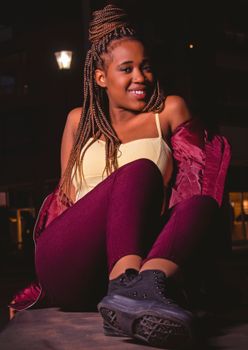 Confident woman with braided hair posing at night in stylish outfit, exuding joy and fashion.