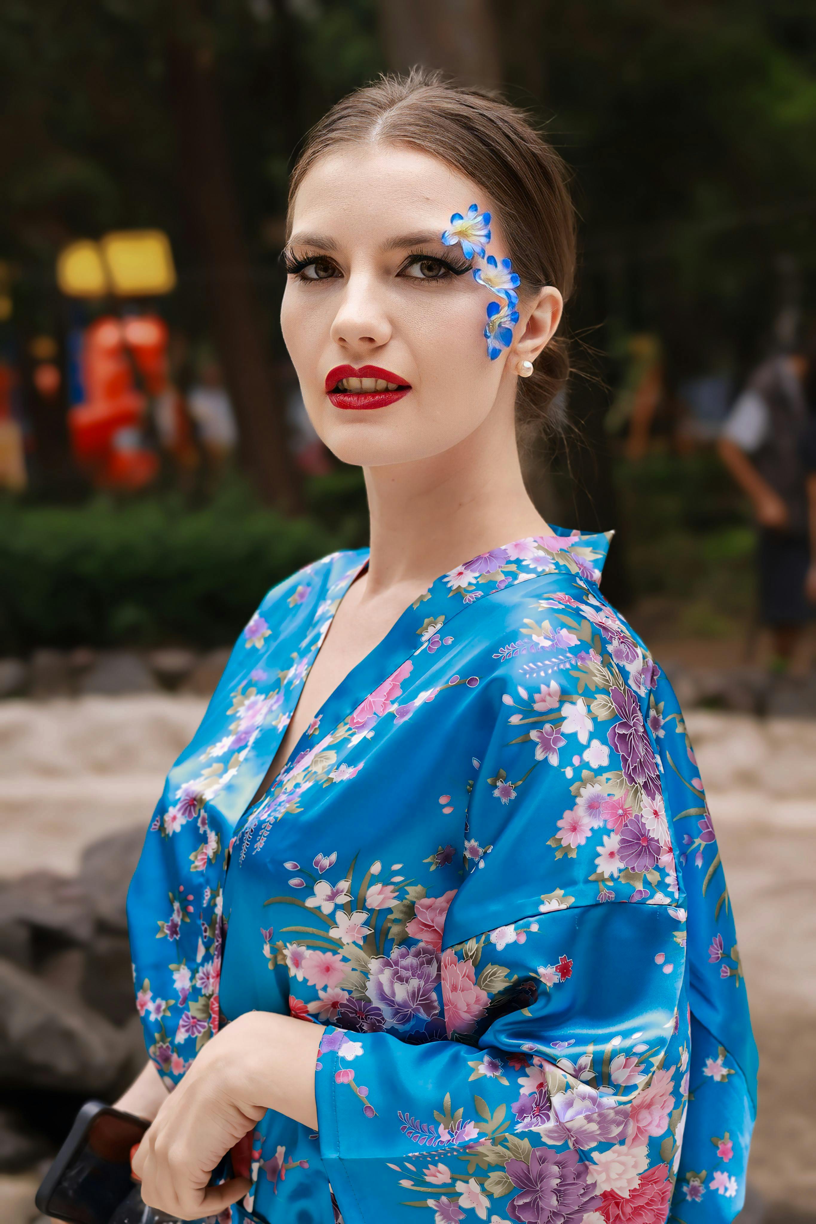 A stylish woman in a blue floral kimono with artistic makeup in an outdoor setting.