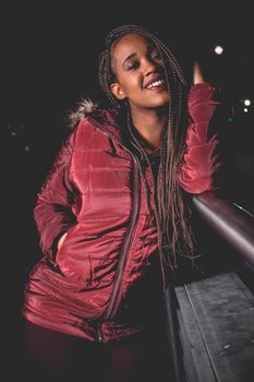 Attractive woman with braided hair in a red jacket smiling outdoors at night.