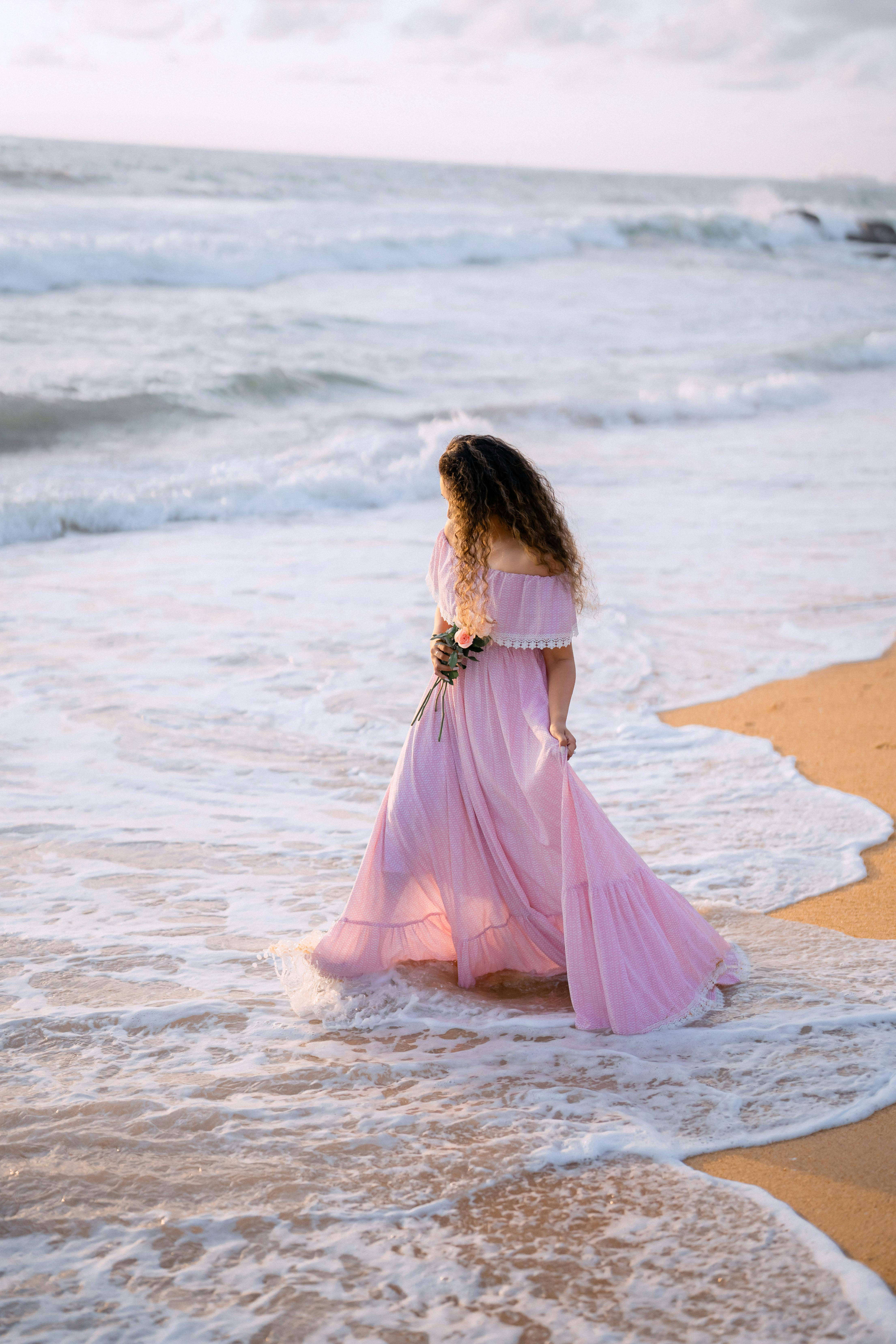 A woman in a pink dress strolls along a serene beach in Sri Lanka, enjoying the ocean breeze.